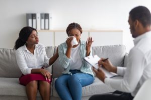 Two black women sitting on a couch in a psychologist's office undergoing counseling with a psychologist and one of the women is crying.