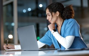 Doctor, laptop and writing notes at night thinking about healthcare solution, idea or planning at hospital. Woman medical nurse working late in focus with notebook and computer for research at clinic.