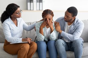 Young African American woman crying at therapist's office, affectionate husband supporting her with professional psychologist. Millennial lady with her spouse on session with marital counselor