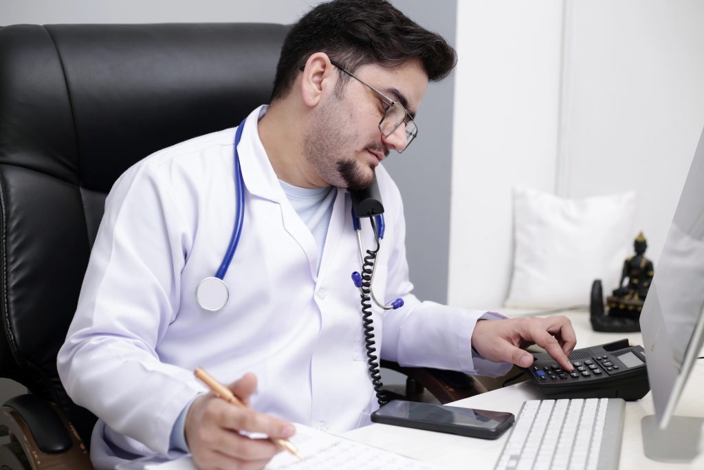 A young doctor is sitting in the clinic and talking on telephone and doing paperwork that should be done by an an admin assistant